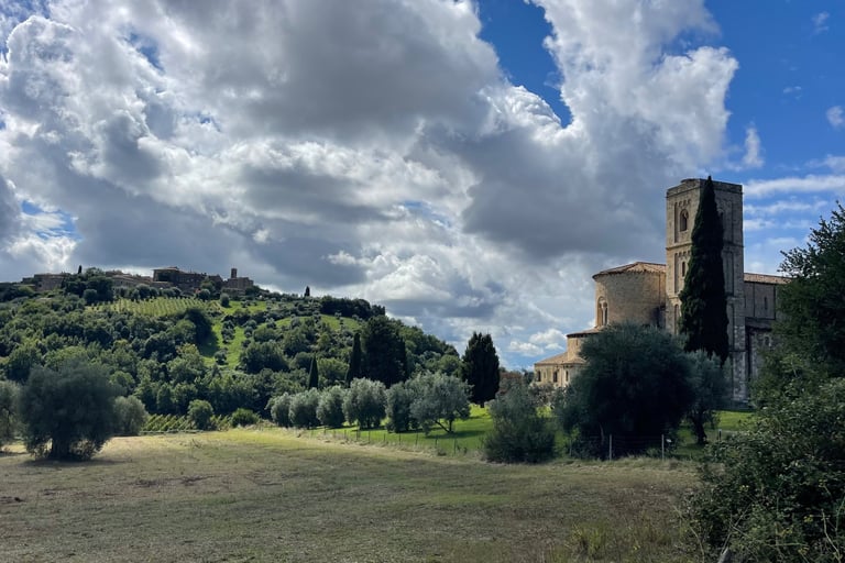 Medieval Abbey of Sant'Antimo in Tuscany surrounded by rolling hills and olive trees under a cloudy blue sky.