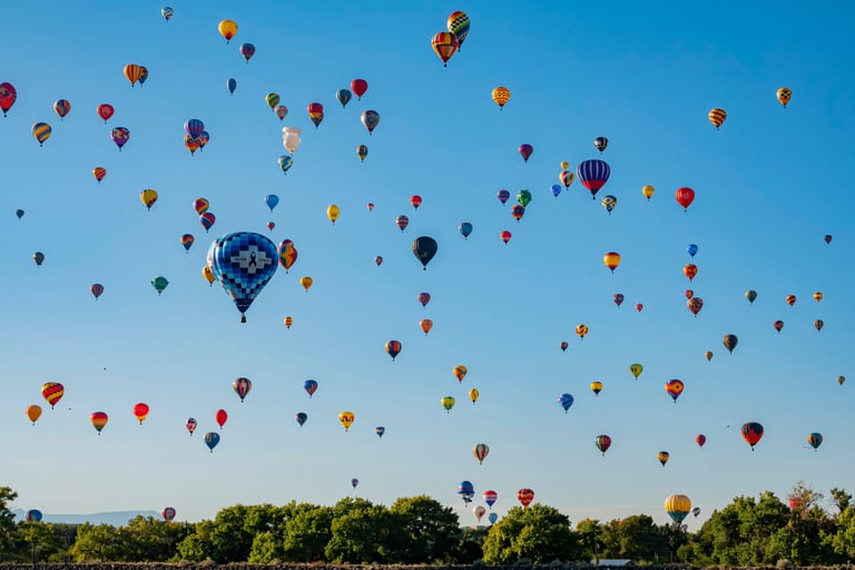 Dozens of colorful hot air balloons floating in a clear blue sky above a line of green trees.