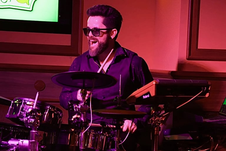 Singer Leo Couto playing the drums, wearing a black shirt, a vest, and sunglasses.