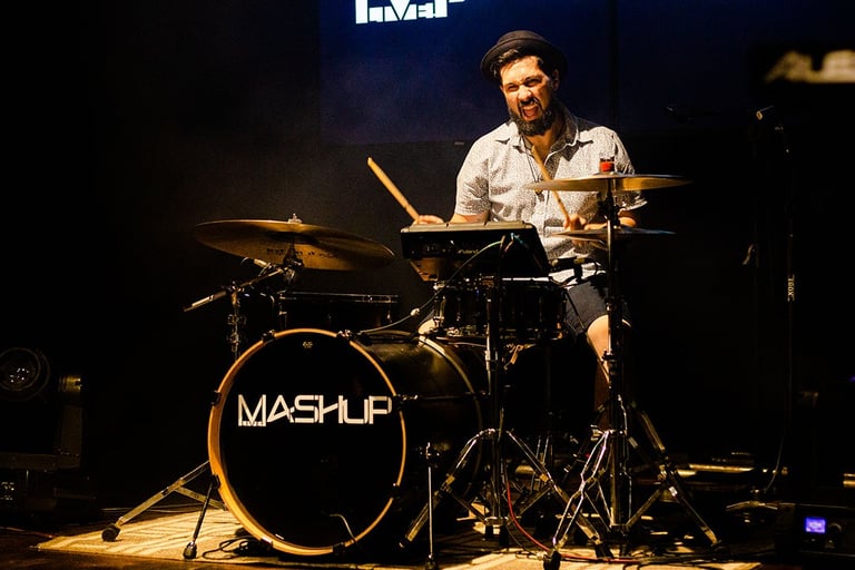 Drummer Lucas Dias wearing a short-sleeved white shirt and a pork pie hat, playing a black drum set.