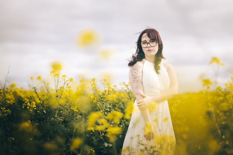 A woman with glasses and a white dress posing in a blooming yellow wildflower field.