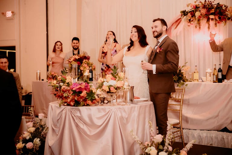 A bride and groom raising a champagne toast at their elegant indoor wedding reception head table.