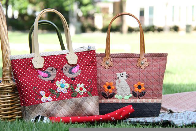 Two quilted fabric tote bags with appliques displayed on grass during a picnic setup.