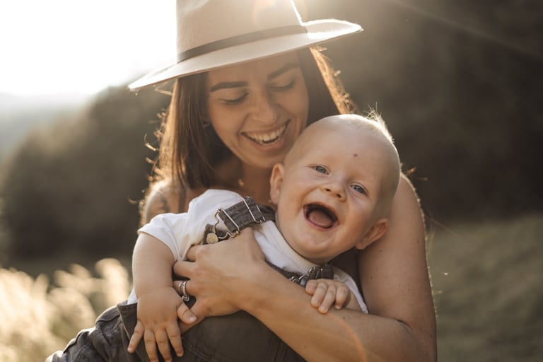 mother holding her baby on the arms and both laughing during sunset 