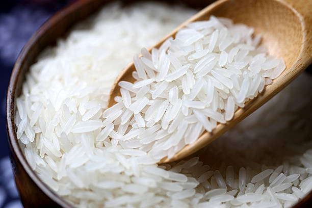 A wooden scoop lifting raw long-grain white jasmine rice from a dark bowl.