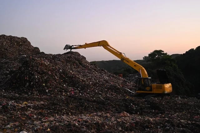 Yellow excavator processing waste at a landfill site during sunset for environmental management.