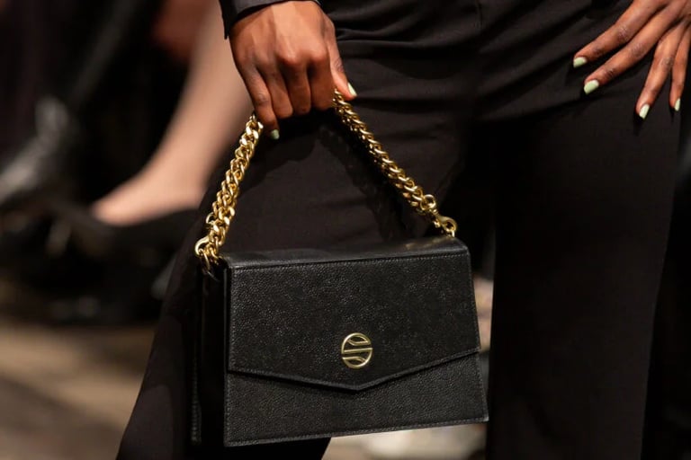 Model holding a black leather designer handbag with a gold chain strap at a fashion show runway.