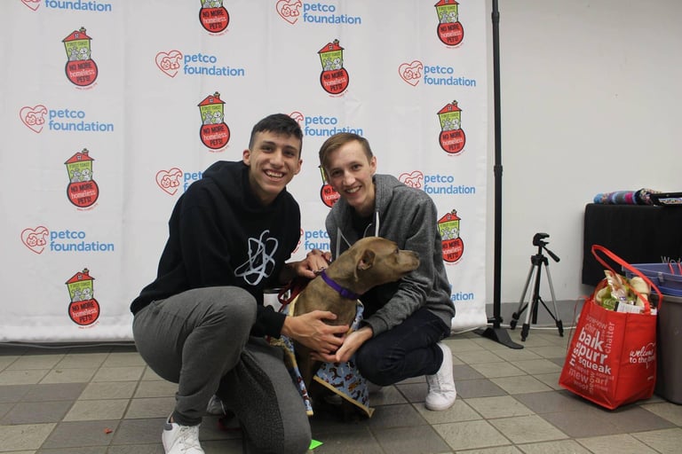 Two smiling individuals crouch beside a brown dog in front of a Petco Foundation backdrop.