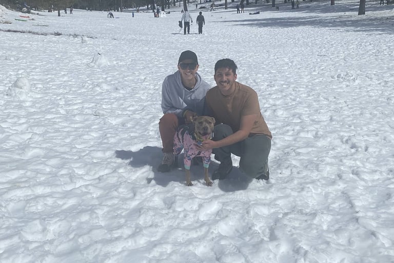 Two men crouched in a snowy field, smiling at the camera with a dog in a pink jacket between them.