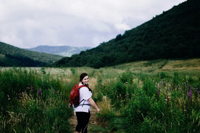 Smiling woman with a red backpack hiking on a scenic mountain trail through a lush green valley.