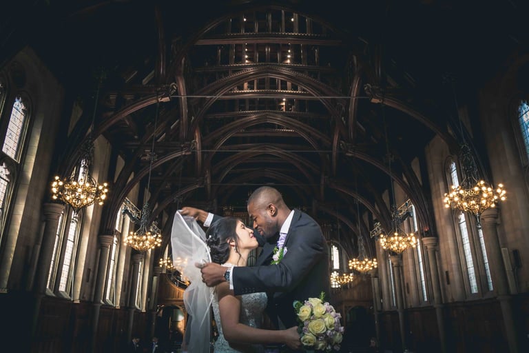 A bride and groom embrace under a veil inside a grand cathedral with vaulted wood ceilings and chandeliers.