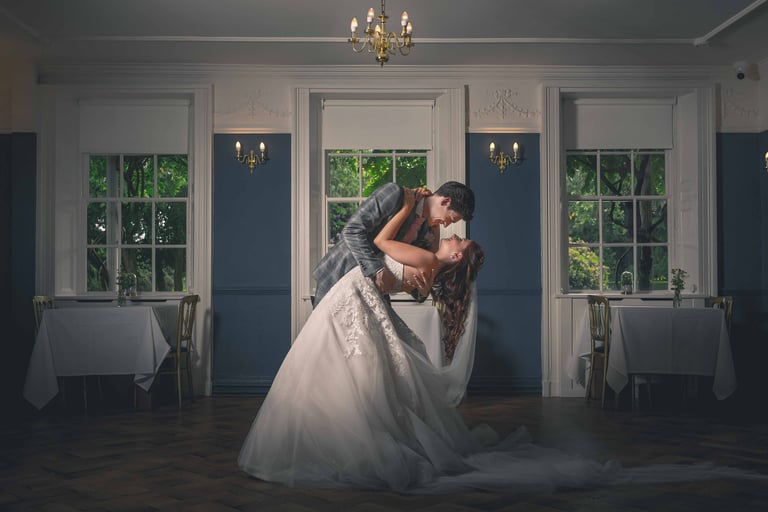 A groom dips his bride during their first dance in an elegant wedding ballroom with blue walls.