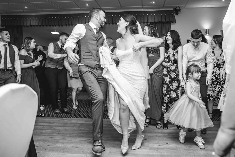 Black and white photo of a happy bride and groom dancing with wedding guests at a reception.