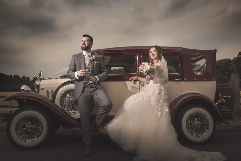 A bride and groom posing for wedding photos next to a vintage burgundy and cream classic car.