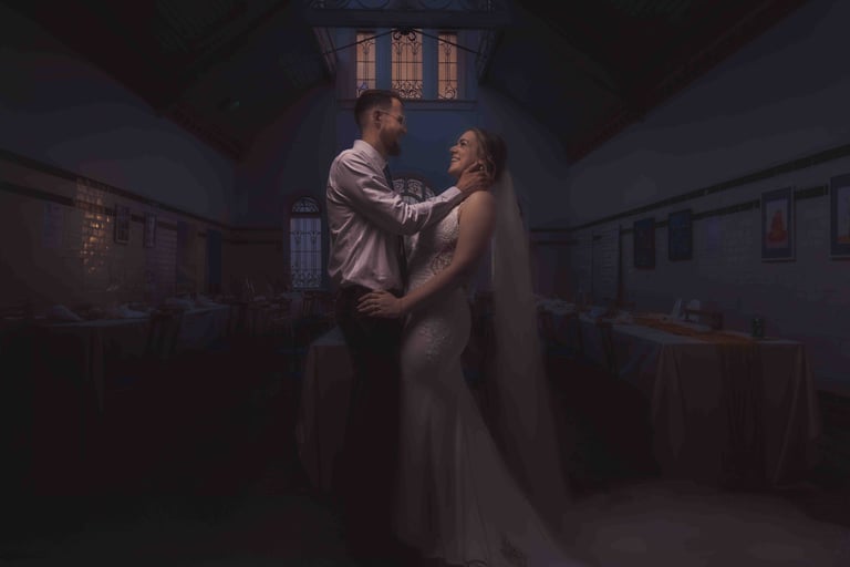 Smiling bride and groom embrace in a dimly lit, historic wedding reception hall with vaulted ceilings.