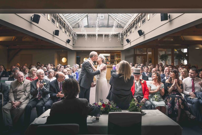 A bride and groom sharing their first kiss during a modern indoor wedding ceremony surrounded by guests.