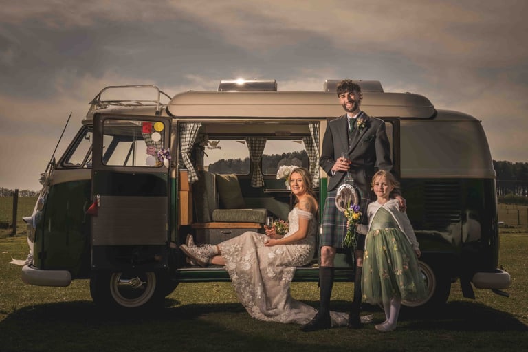 Scottish wedding couple in a kilt and lace gown posing with a child by a vintage VW camper van.