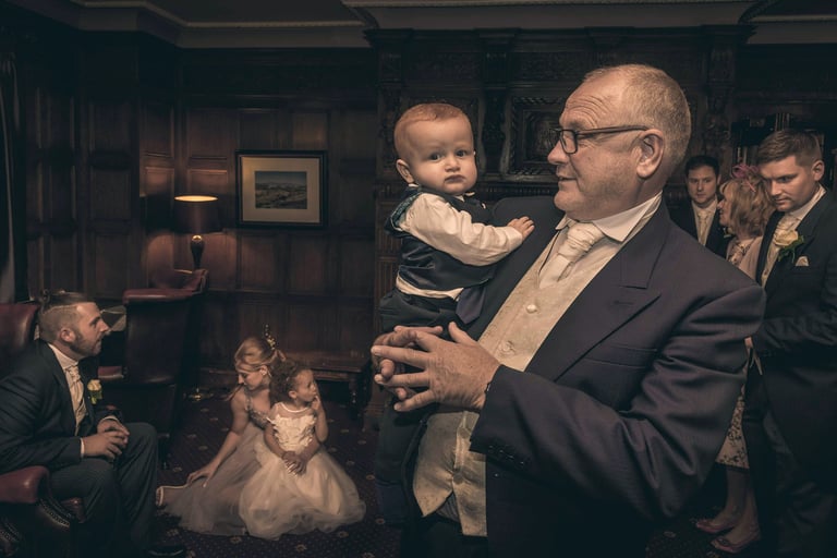 Grandfather in a formal suit holding a baby boy at a wedding reception in a wood-paneled room.