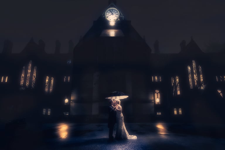 A bride and groom sharing a romantic kiss under an umbrella at night in front of a gothic clock tower.