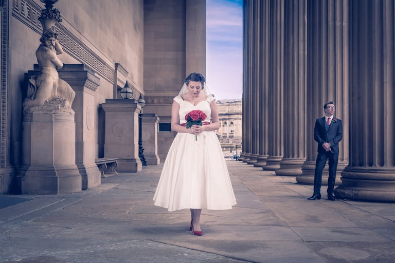 A bride in a vintage tea-length wedding dress holds red roses outside a neoclassical building with columns.
