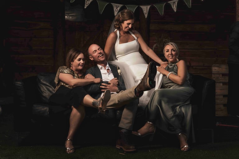Smiling bride and wedding guests laughing together on a black leather sofa at a rustic reception venue.
