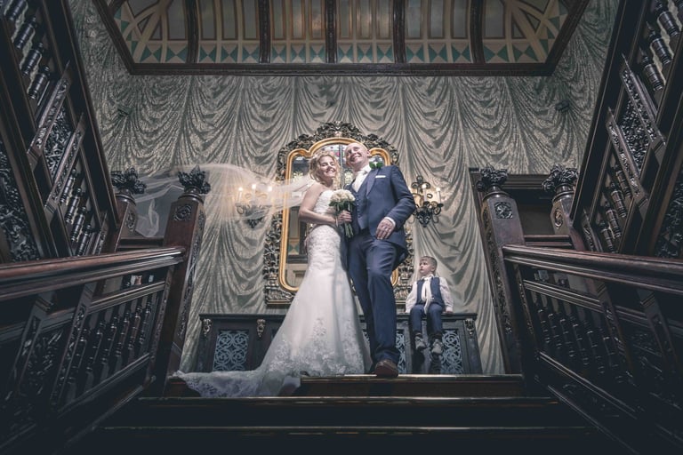 Smiling bride and groom posing on a grand wooden staircase in a luxury wedding venue.