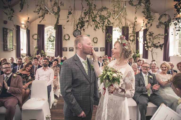 Smiling bride in floral crown and groom at an indoor rustic wedding ceremony with greenery decor.