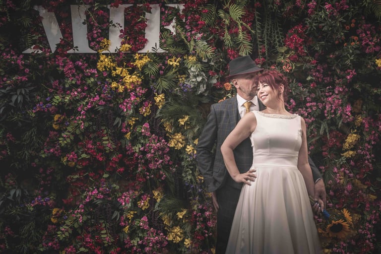 A smiling bride and groom pose in front of a colorful flower wall at a wedding venue.