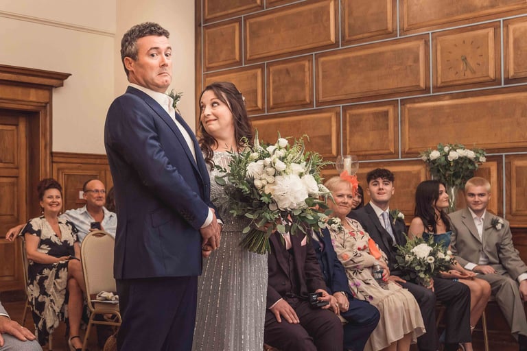 A groom making a funny face during a wedding ceremony in a wood-paneled room while the bride looks on.