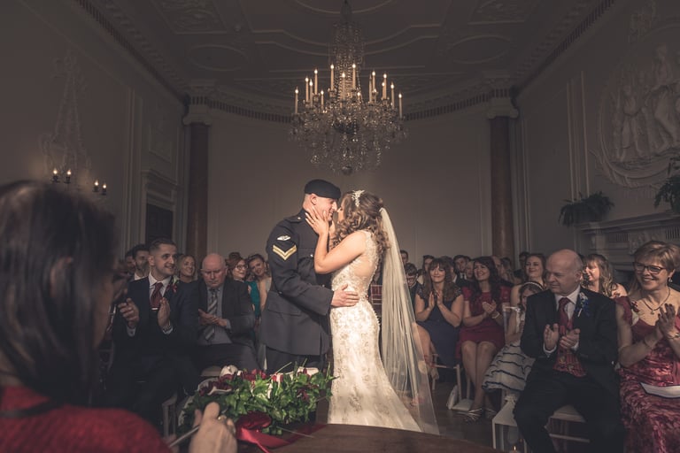 A military groom in uniform kisses his bride in a lace wedding dress at a luxury indoor ceremony.