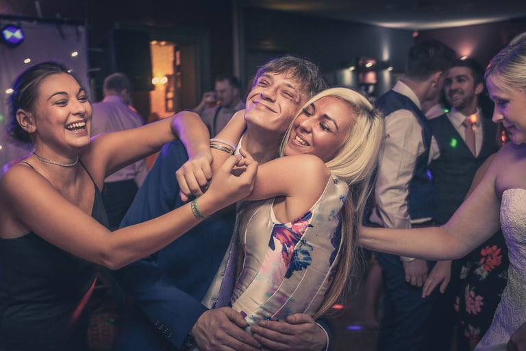 Friends laughing and hugging on a dance floor at a wedding reception party with club lighting.