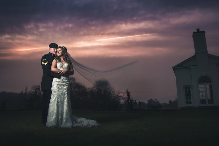 Military wedding couple embracing at sunset with a flowing bridal veil and manor house background.