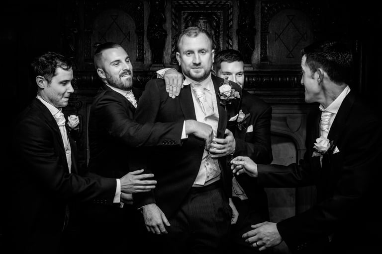 Black and white photo of a groom and four groomsmen in formal wedding suits adjusting his jacket.