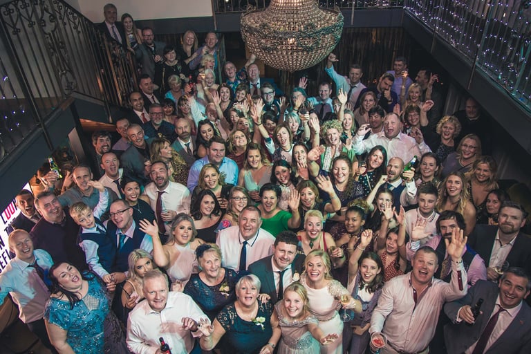 A large group of wedding guests smiling and waving at a party under a crystal chandelier.