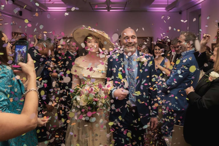 Smiling bride and groom celebrate with guests throwing colorful confetti at an indoor wedding reception.