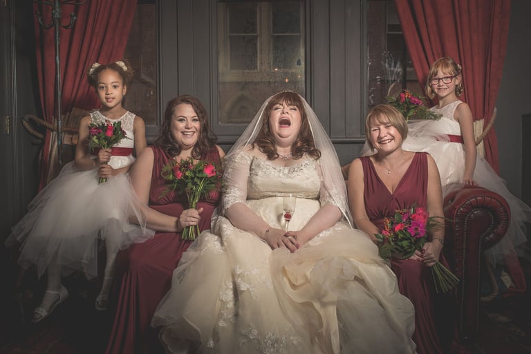 A laughing bride in a lace gown sits with bridesmaids in burgundy dresses and flower girls holding bouquets.