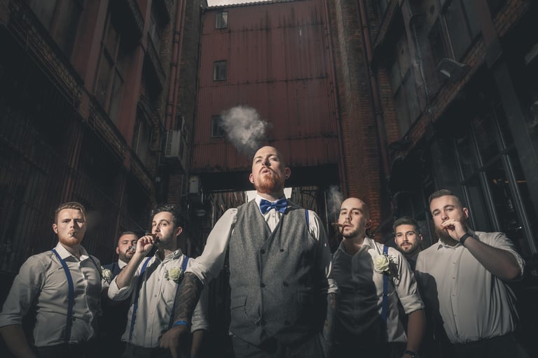Groom and groomsmen in vintage wedding attire smoking cigars in an industrial alleyway.