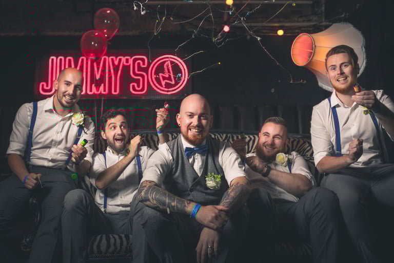 A group of happy groomsmen in suspenders celebrate at a bachelor party in a bar with confetti.
