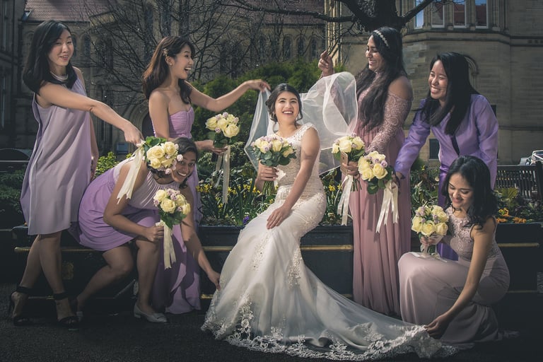 Smiling bride in a white lace gown surrounded by bridesmaids in lavender dresses holding white rose bouquets.