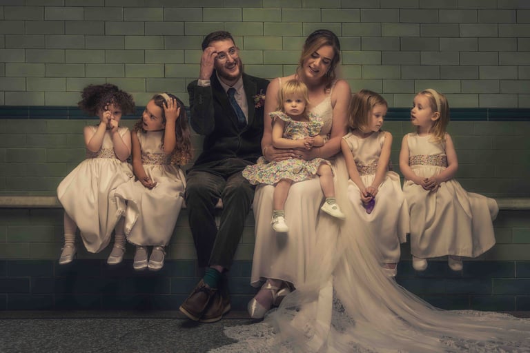 A bride and groom in wedding attire sitting with five flower girls in white dresses against a green tiled wall.