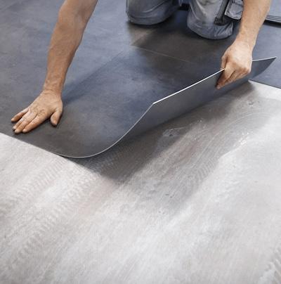 Professional flooring installer laying dark gray vinyl floor tiles over a gray concrete subfloor.