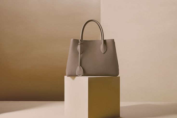 Elegante bolso de mano de piel color topo exhibido sobre un pedestal minimalista.