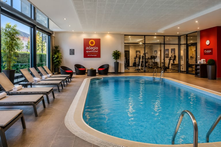 Indoor swimming pool with lounge chairs and fitness center at Adagio Aparthotel Val d'Europe.