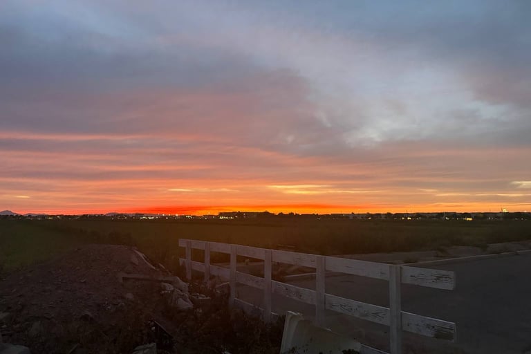 A vibrant orange and purple sunset over a dark field with a white wooden fence in the foreground.