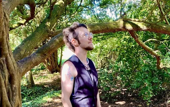 William Elisabeth Cuthbert, a white trans masc person, with trees in the background