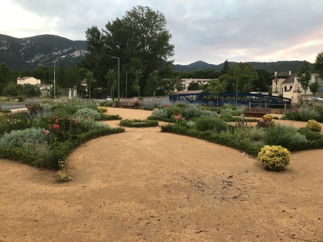 A scenic garden park with sandy paths, lush flowering plants, a blue bridge near Quillan mountains.