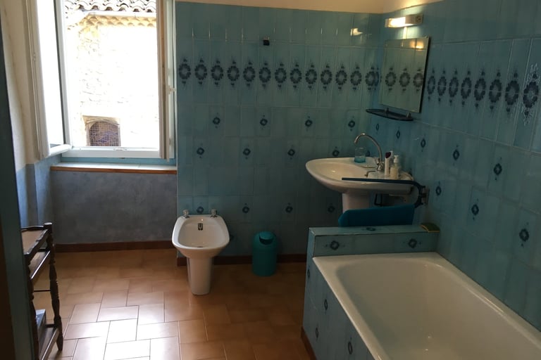 Rustic bathroom featuring blue patterned wall tiles, a white bathtub, bidet, pedestal sink, and terracotta flooring.