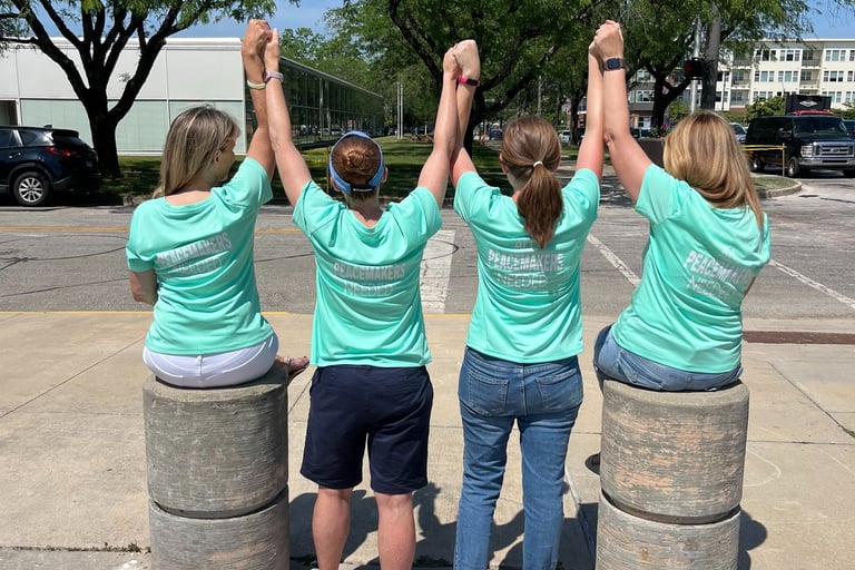 Four people in teal Peacemakers Needed shirts holding hands and raising arms on a sidewalk with chalk art.