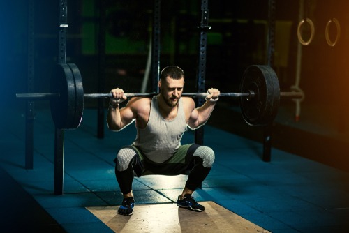 A muscular man performing a heavy barbell back squat in a dark gym for strength training.