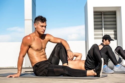 Athletic shirtless man performing core strengthening exercises on a yoga mat during an outdoor fitness class.